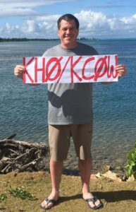 Tom Callas, KC0W, drží ručne napísanú tabuľu s volacím znakom KHØ/KCØW na pobreží Saipanu s výhľadom na tyrkysovú lagunu Mariánskych ostrovov